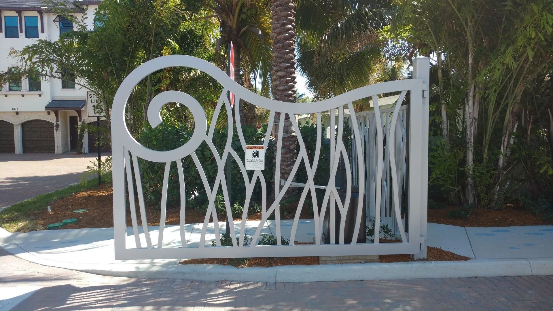 White decorative metal fence with spiral design in front of tropical landscaping and a building.