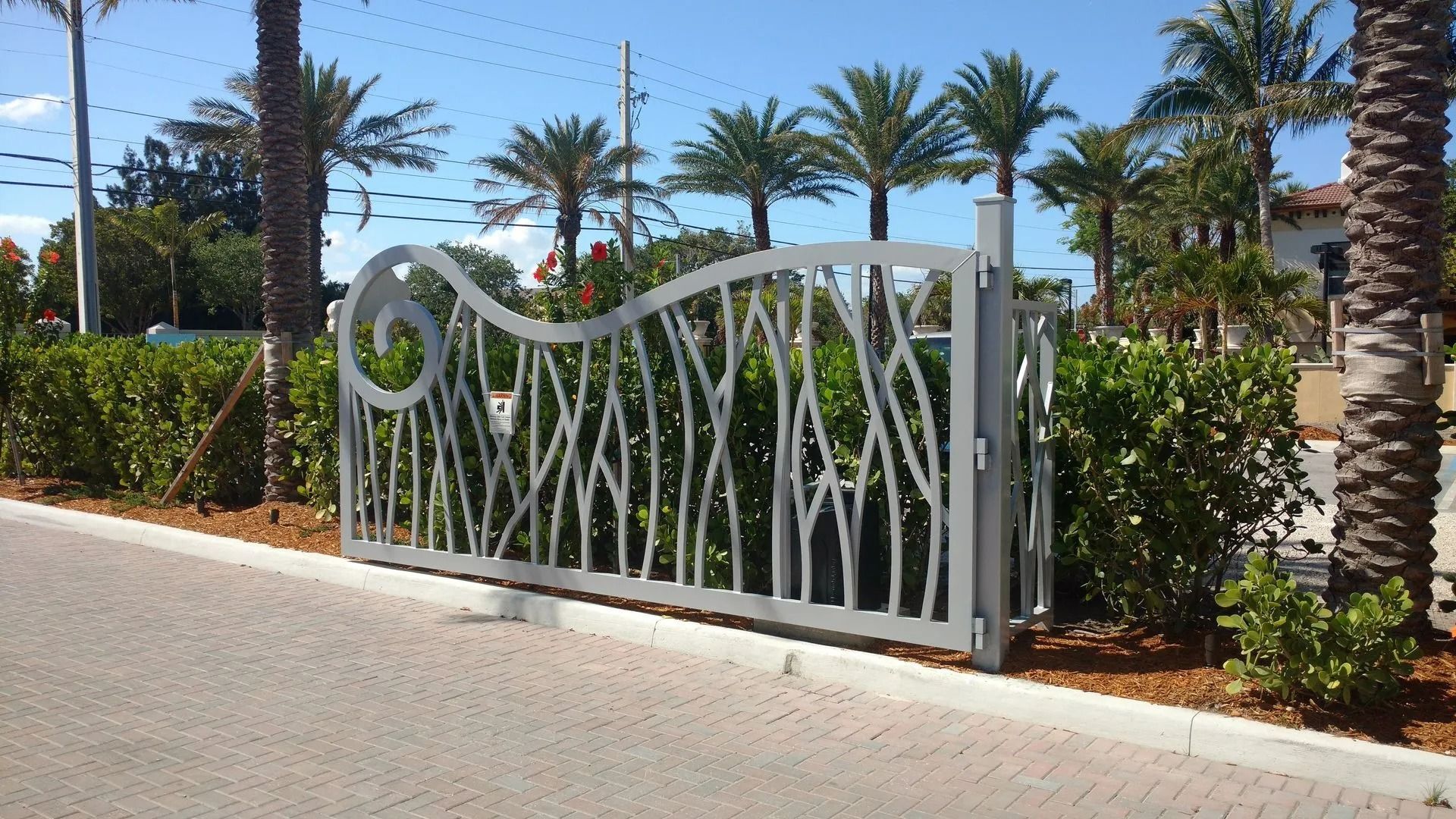 White decorative gate at a landscaped roadside entrance with palm trees and shrubs behind it