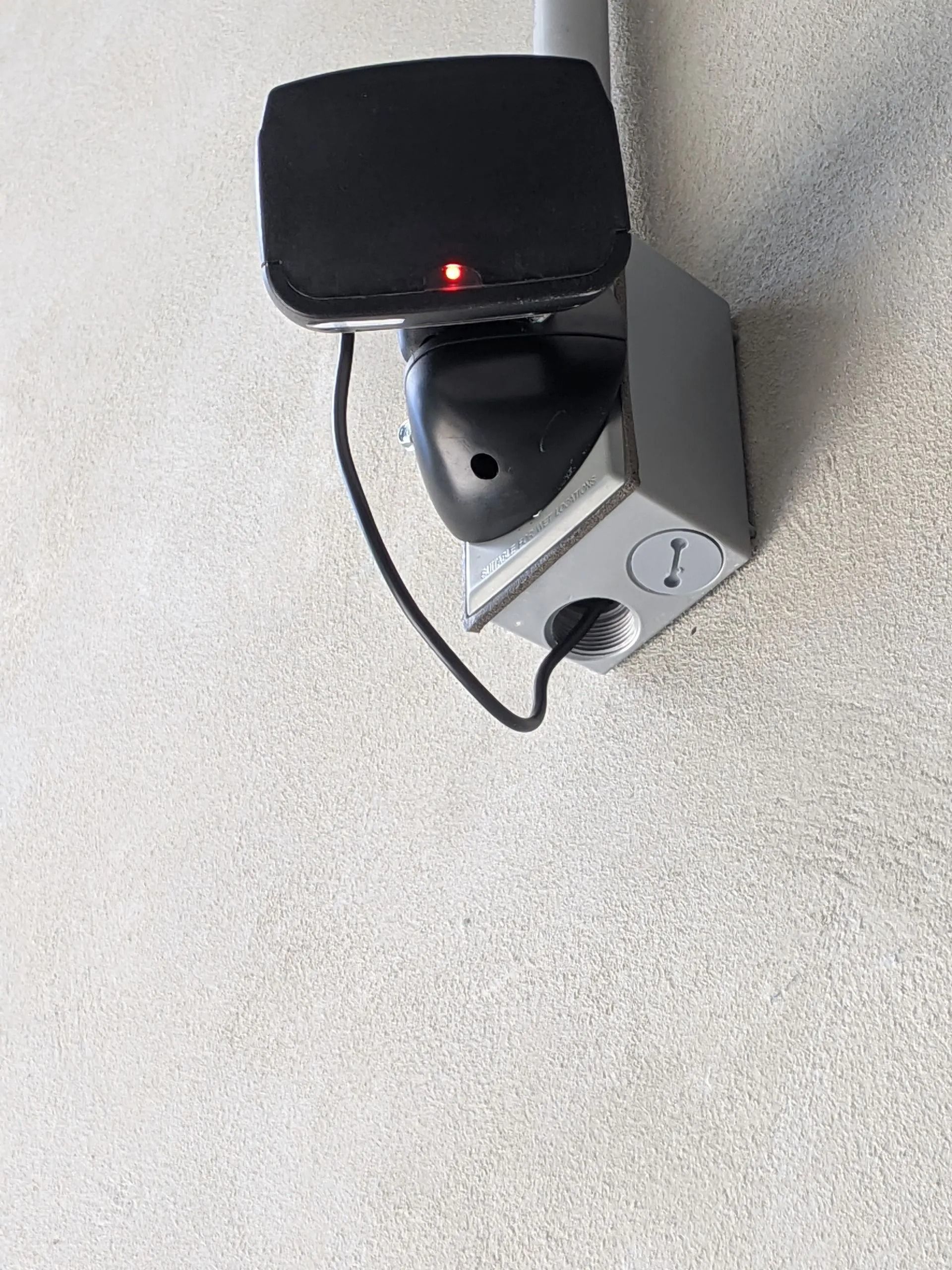 Wall-mounted black device with a red light above a gray box and hanging cable on a textured wall