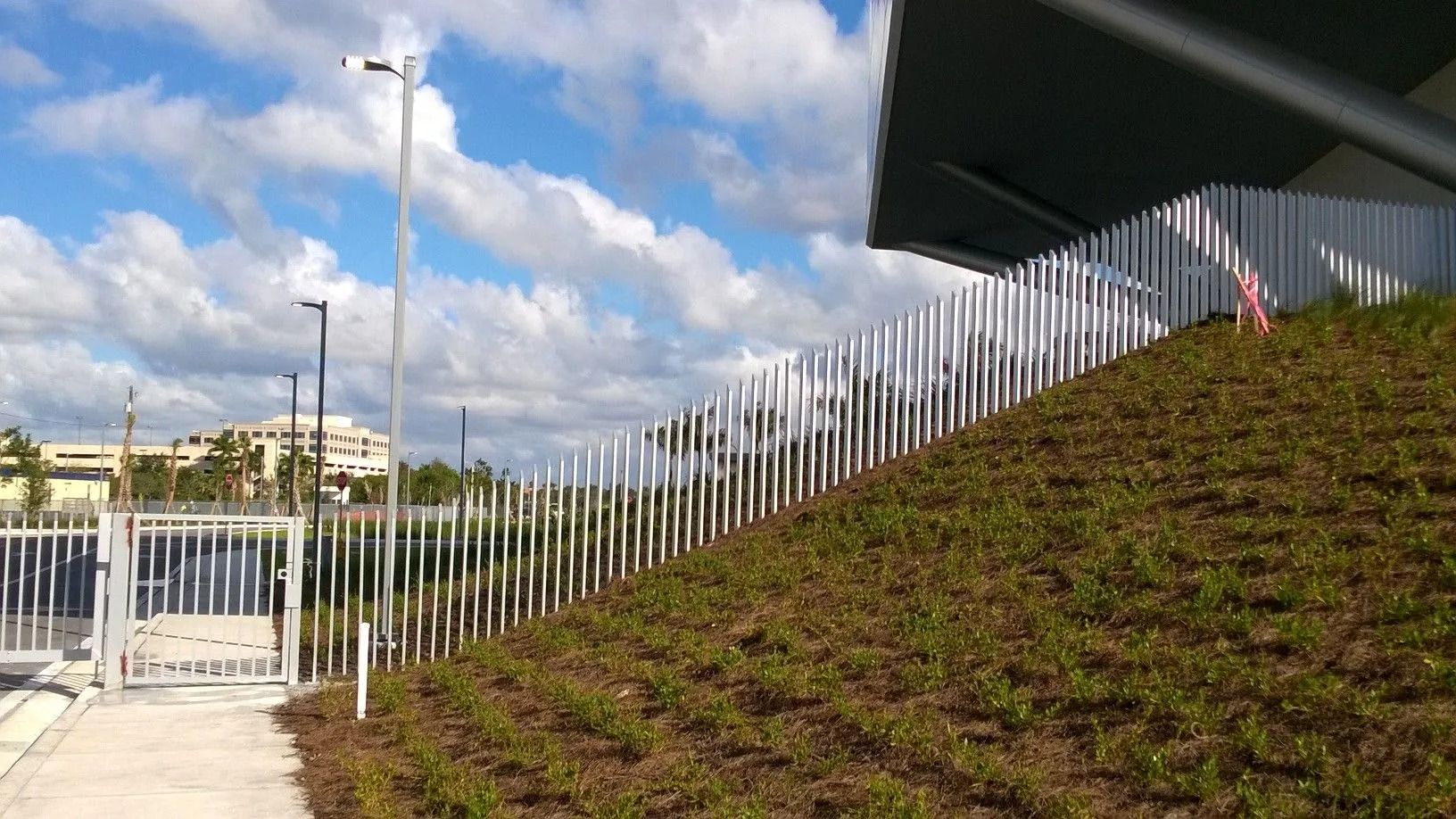 Walkway beside a grassy embankment with metal fence and a building under a cloudy sky