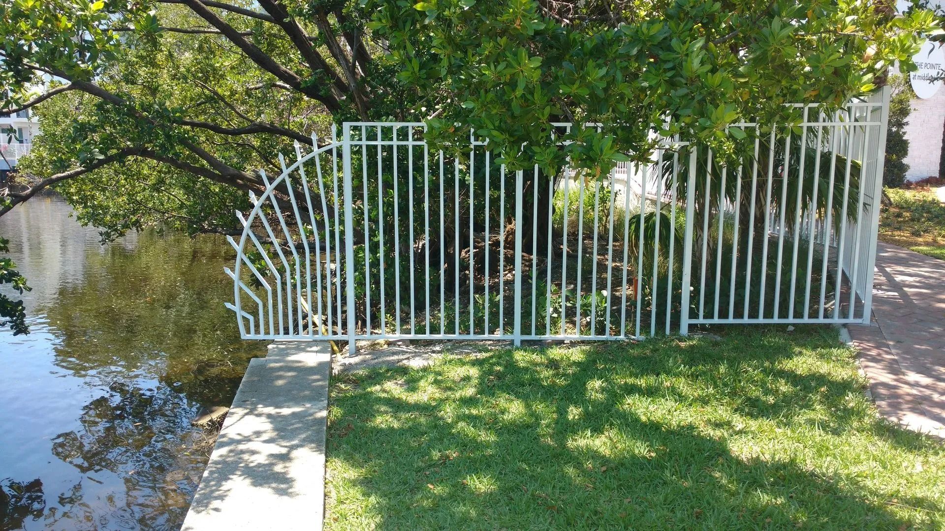 White metal gate beside a grassy yard and sidewalk under trees by a calm waterway