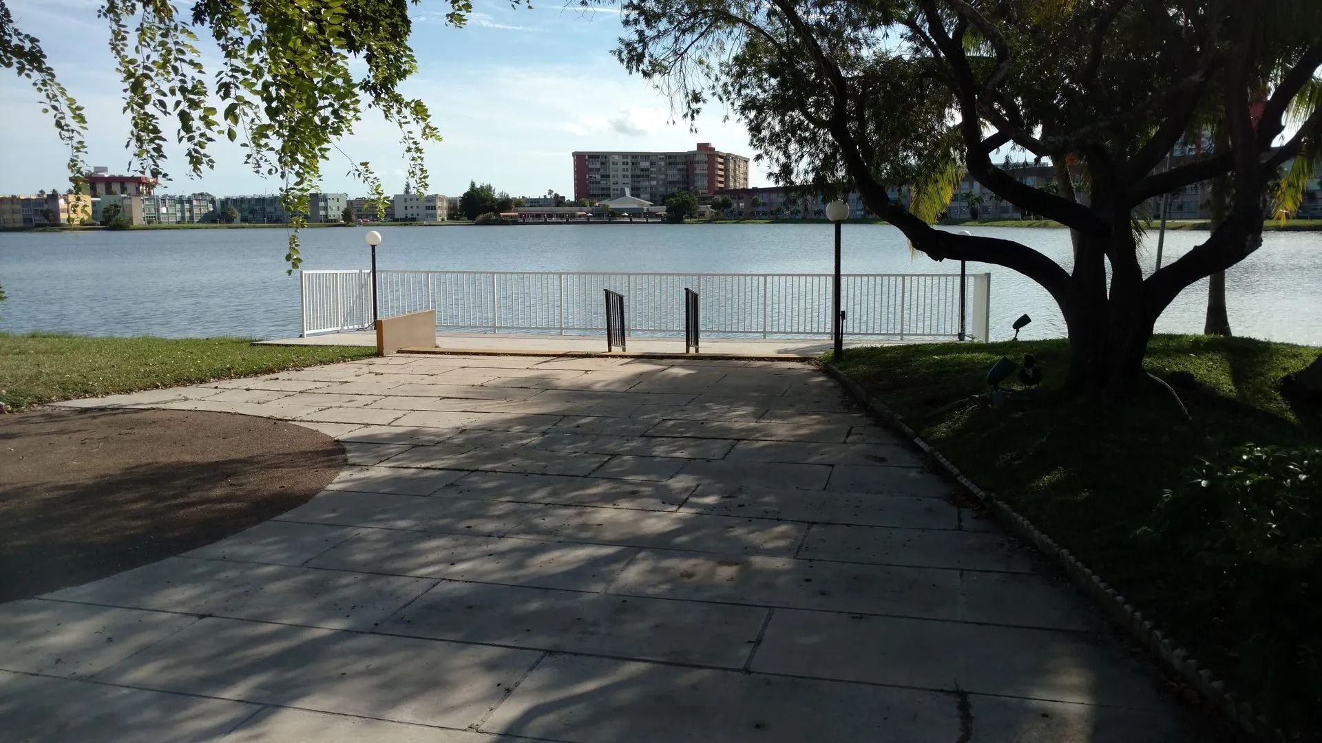 Shaded paved path leading to a fenced waterfront with trees and buildings across the water