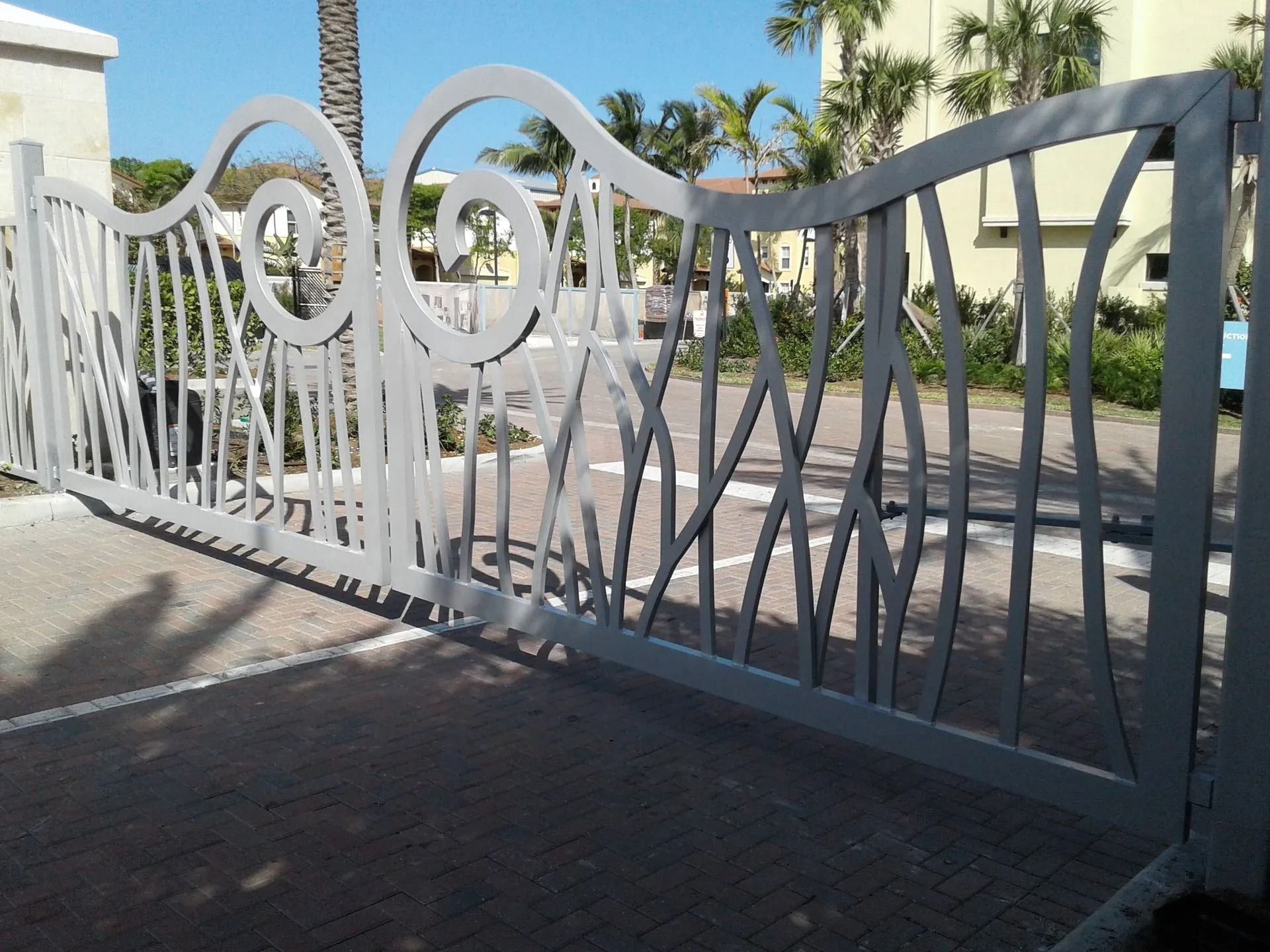 White decorative metal gate with spiral and wavy patterns across a driveway in sunlight