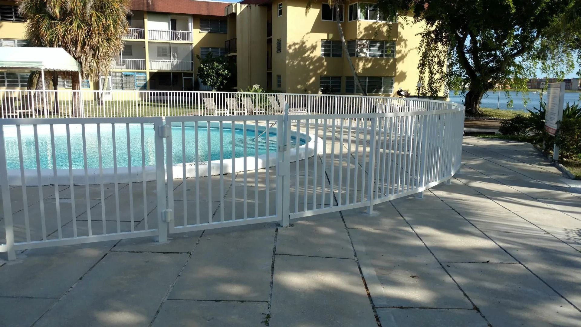 Swimming pool behind a white fence in an apartment courtyard with trees and paved walkway