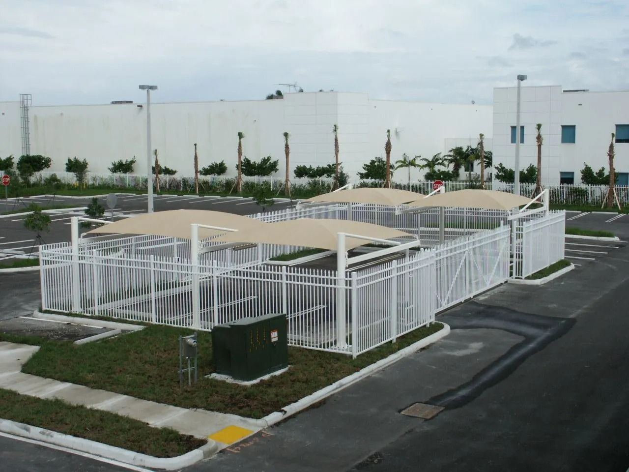 White fenced utility enclosure beside a road, with paved paths and a white building in the background.