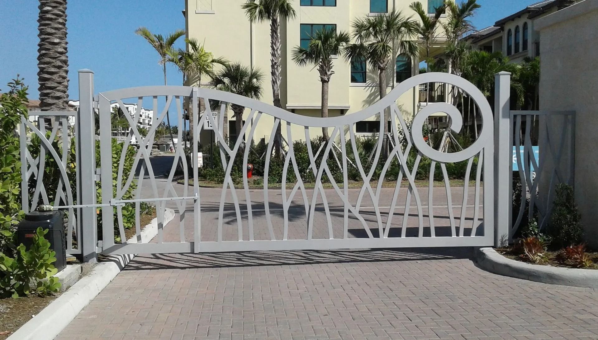 White gated driveway entrance in front of a beige building and palm trees