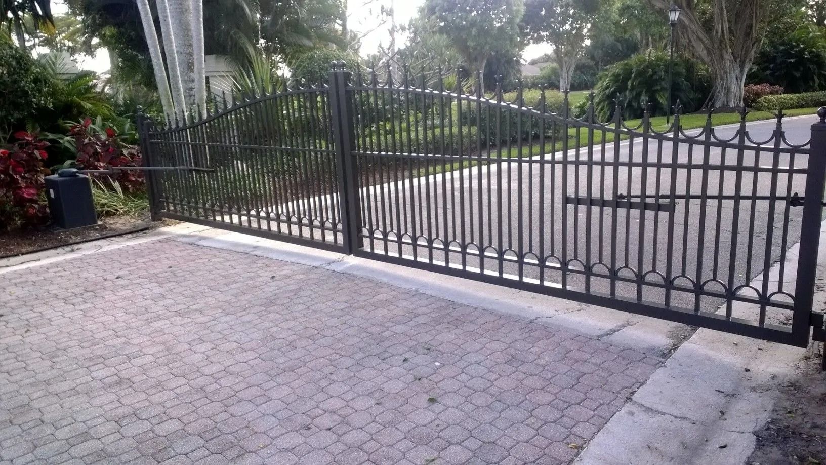 Black metal driveway gate beside a paved entry, with trees and a road in the background.