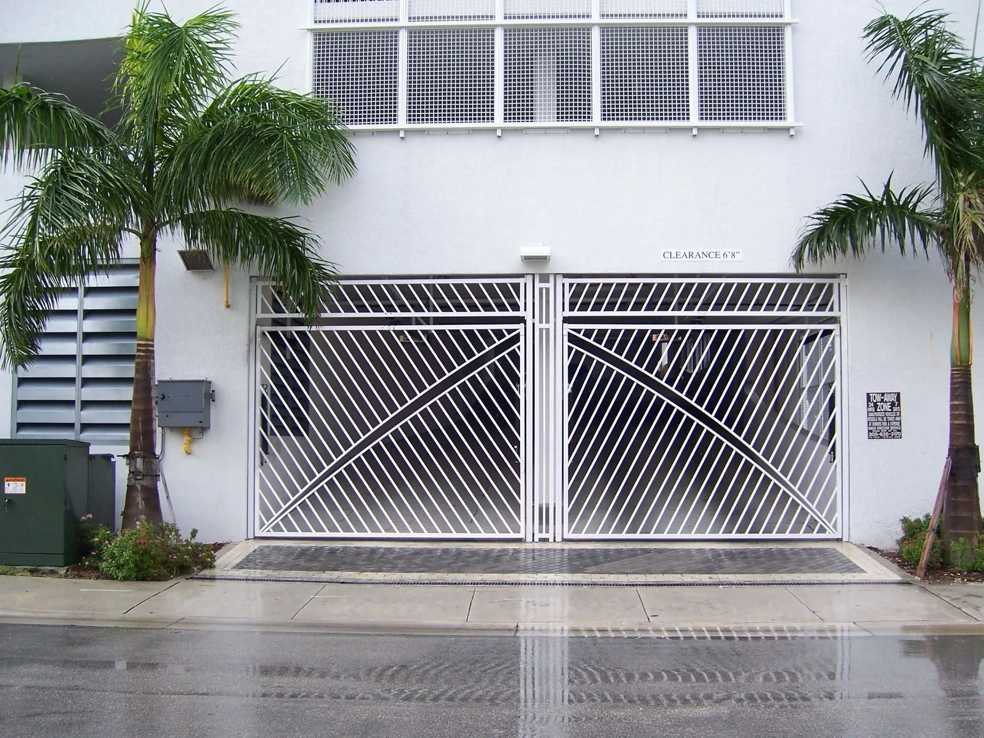 White house facade with large metal gate, palm trees, and wet driveway.