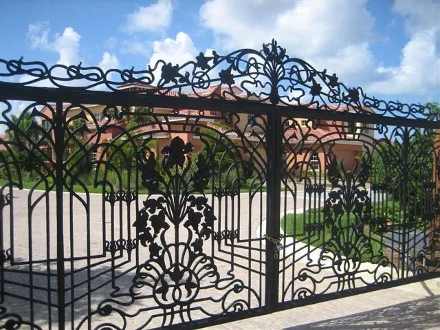 Ornate black wrought-iron gate in front of a residential courtyard with brick buildings and greenery