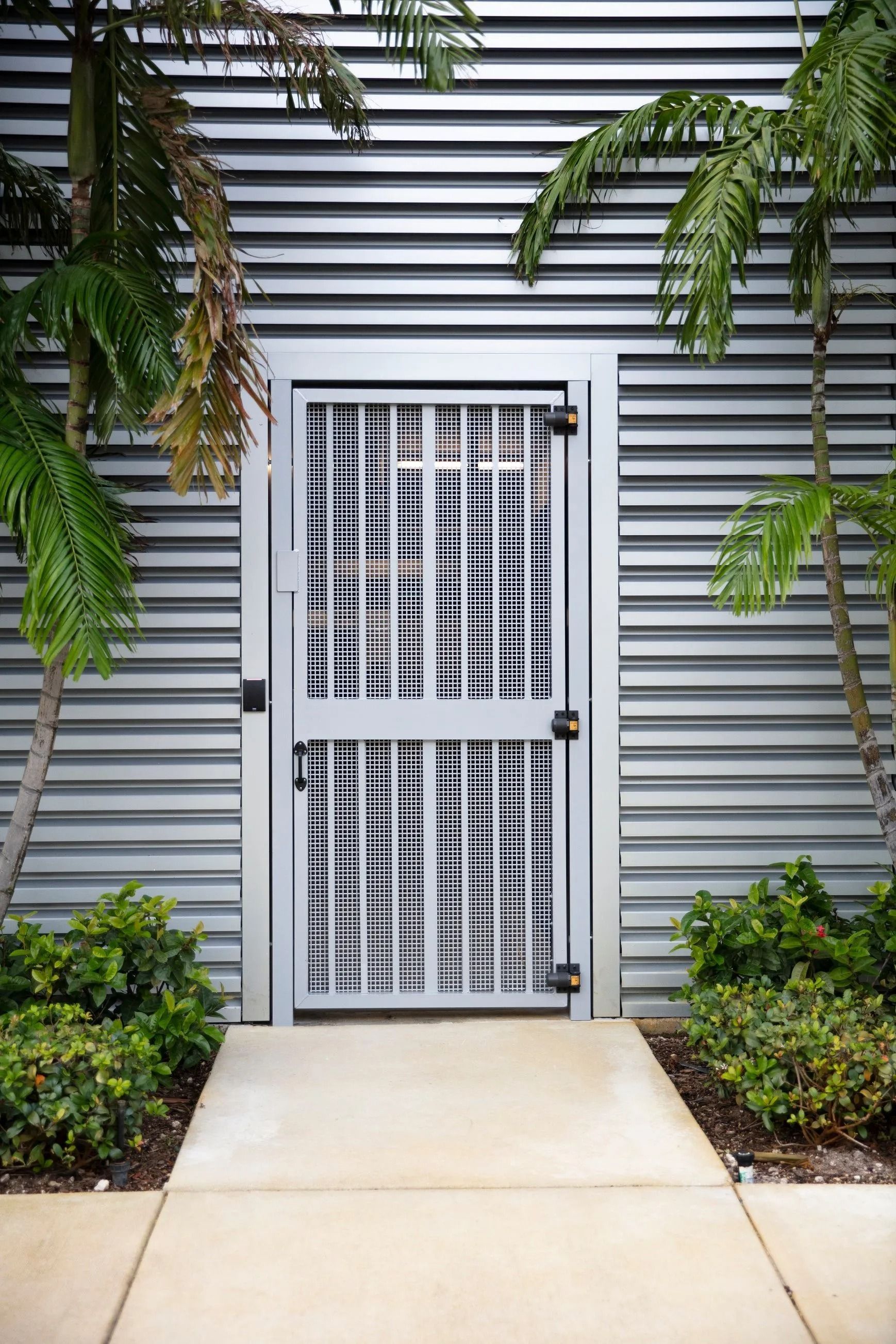 White metal security door in a gray siding wall, framed by green plants and a concrete walkway