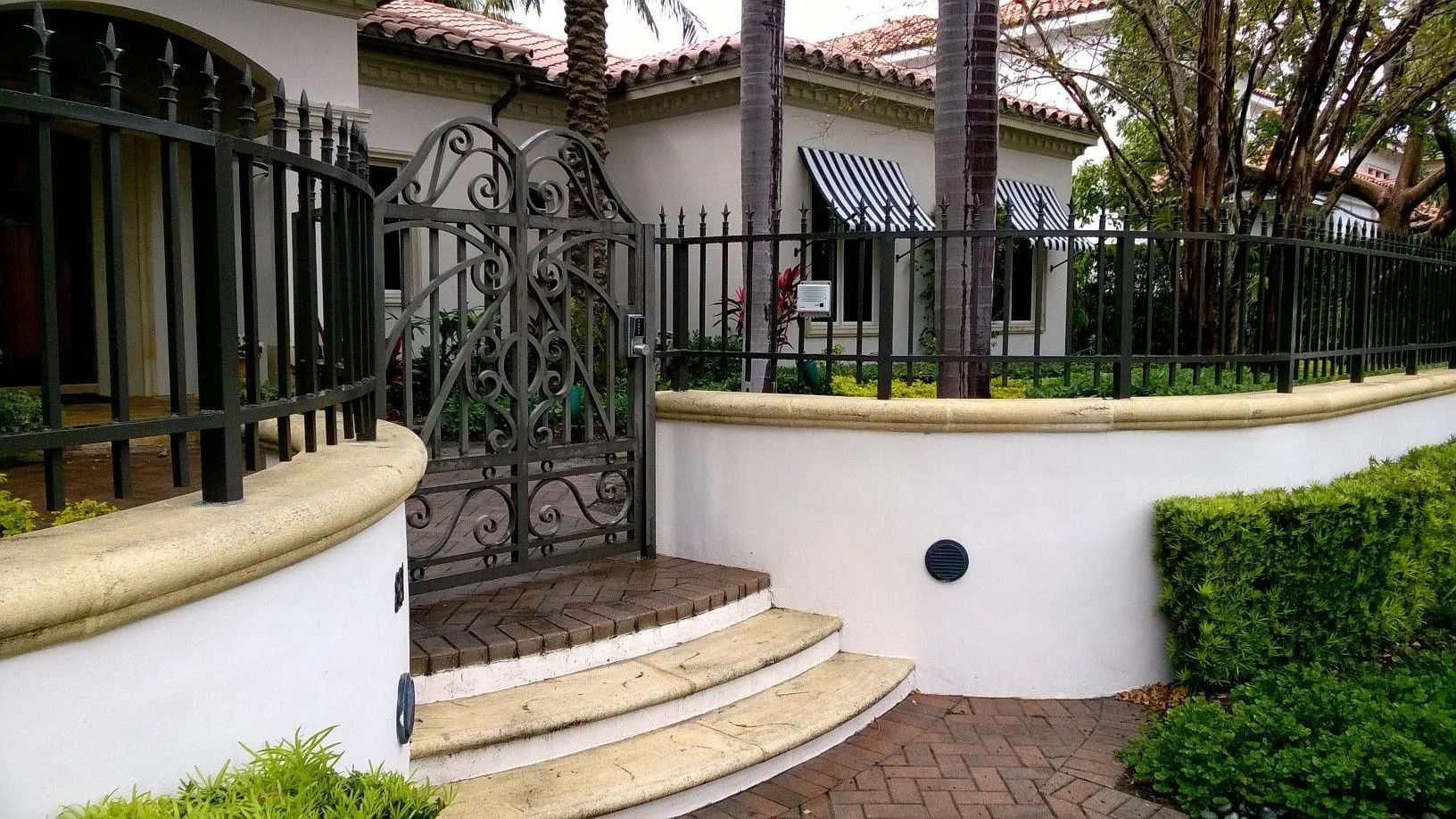 Curved white wall and steps leading to a wrought-iron gate at a house entrance with palm trees.