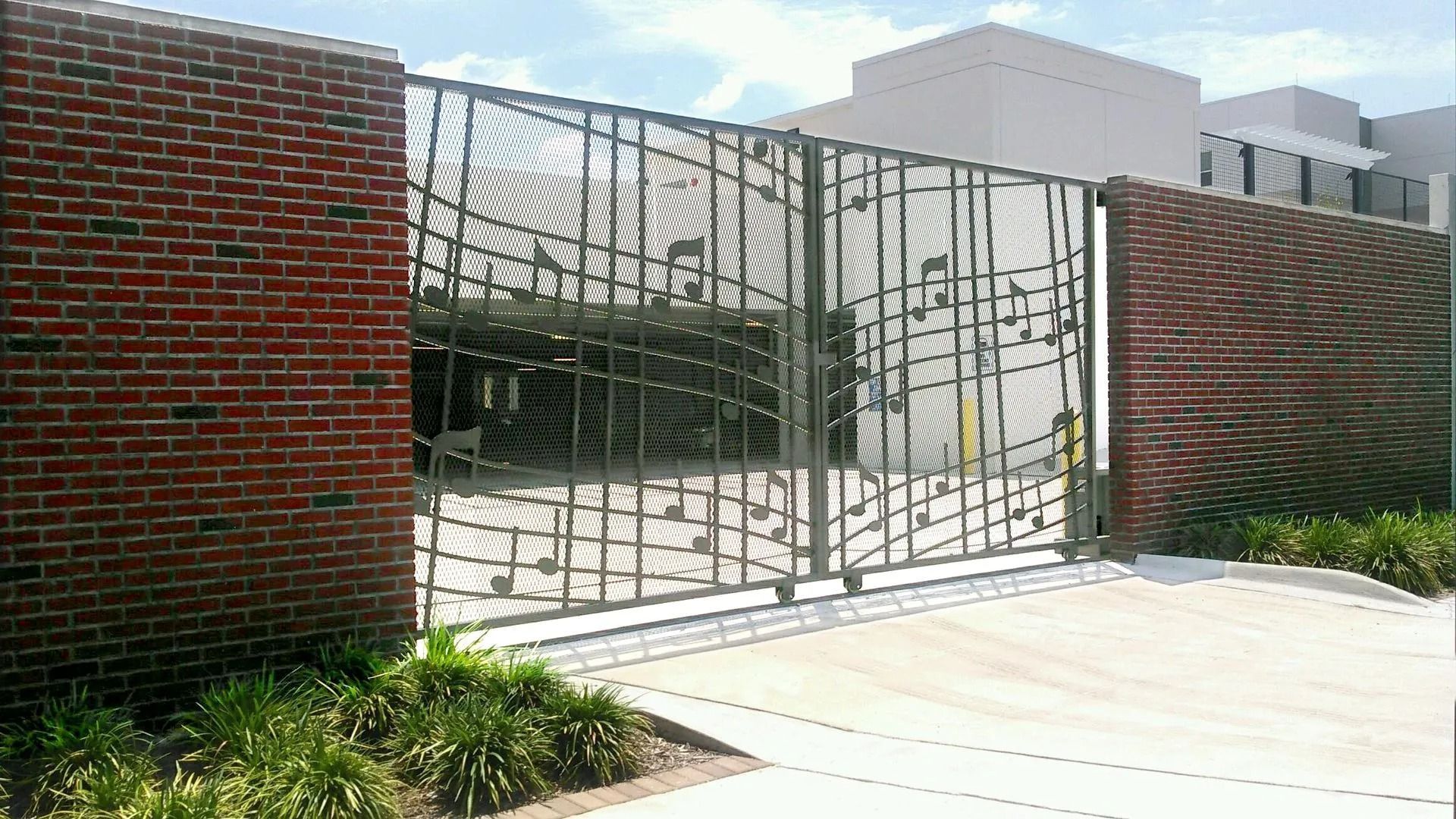 Brick wall and metal gate entrance to a parking area, with sunlight and landscaping.