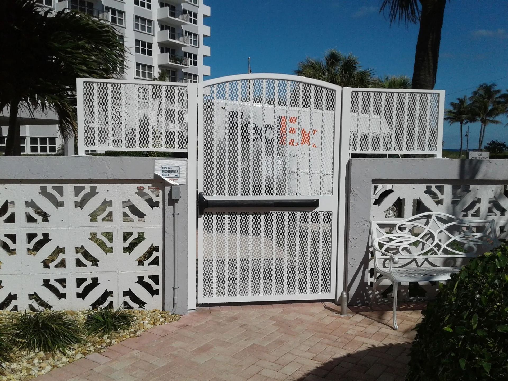 White decorative gate and fence at a building entrance with a brick walkway and palm trees