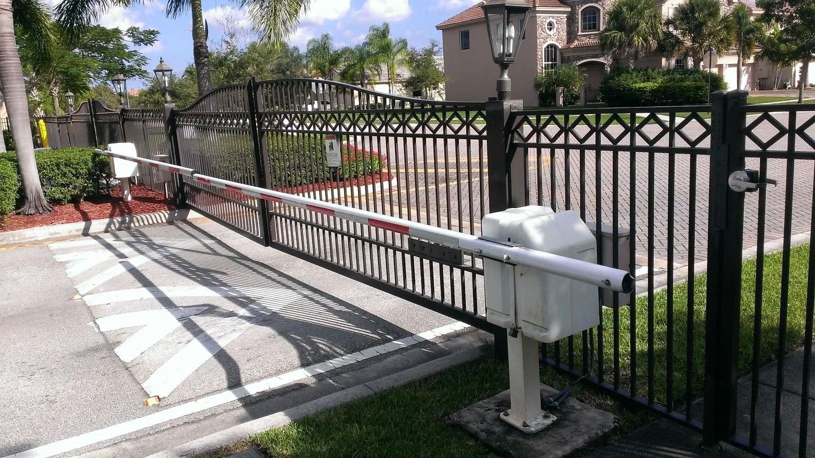 Driveway access gate with a white barrier arm beside a black metal fence and sidewalk ramp.