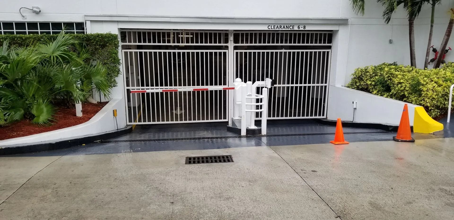 Building entrance blocked by white metal gates, orange cones, and red-white caution tape in a driveway.