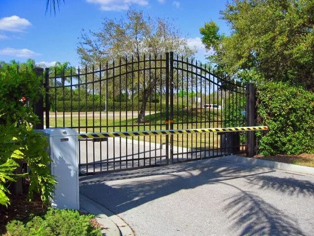 Black wrought-iron driveway gate at a landscaped entrance, with trees and blue sky behind.