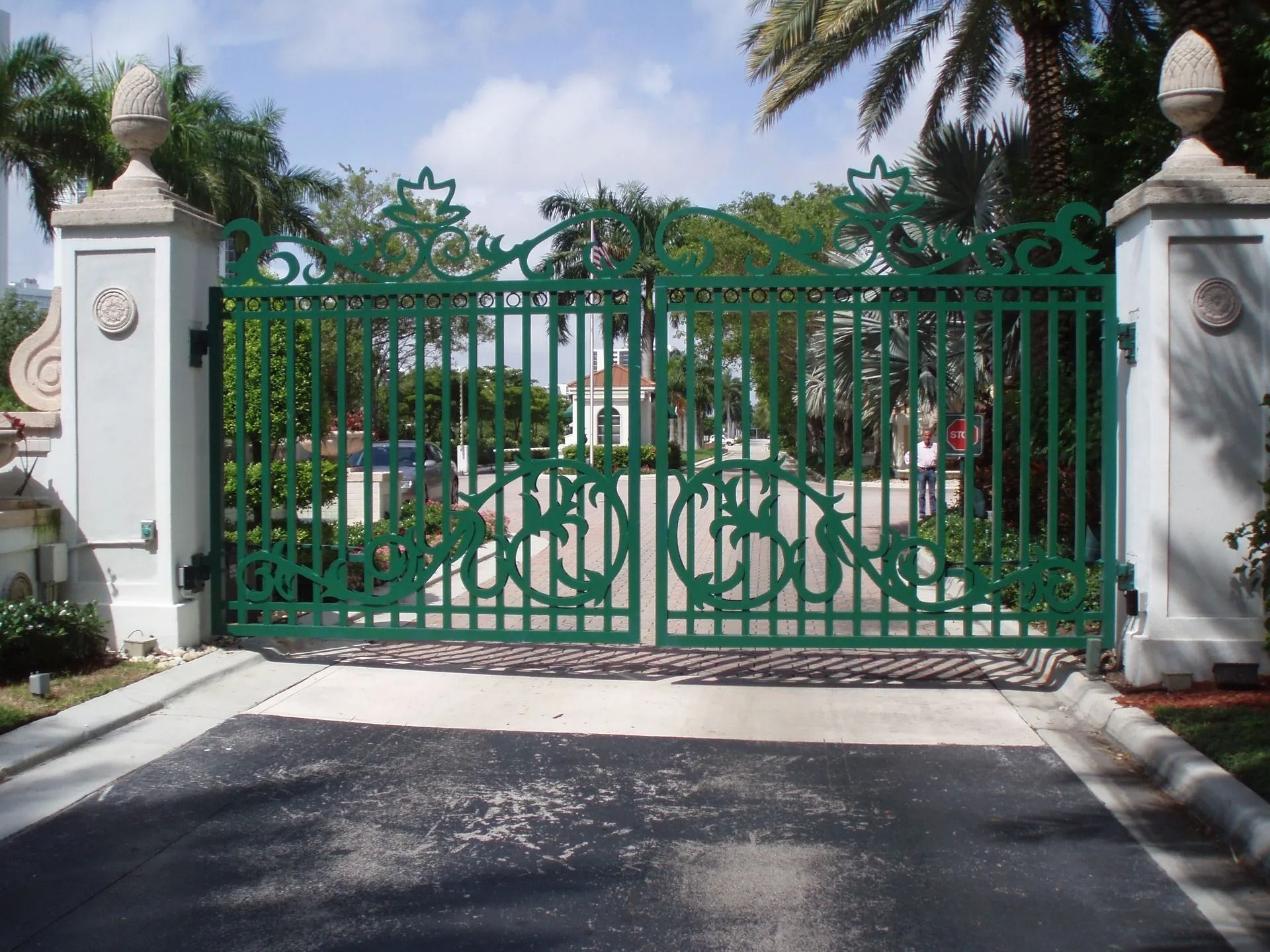 Green ornate gated driveway entrance between white pillars with palm trees behind it