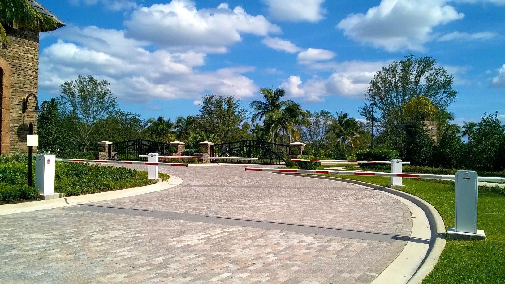 Curving cobblestone driveway lined with white posts, green trees, and a blue sky with clouds