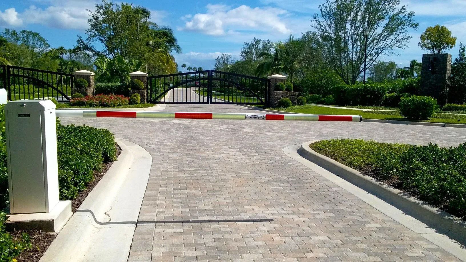 Brick driveway leading to a closed black gate in a sunny landscaped entrance