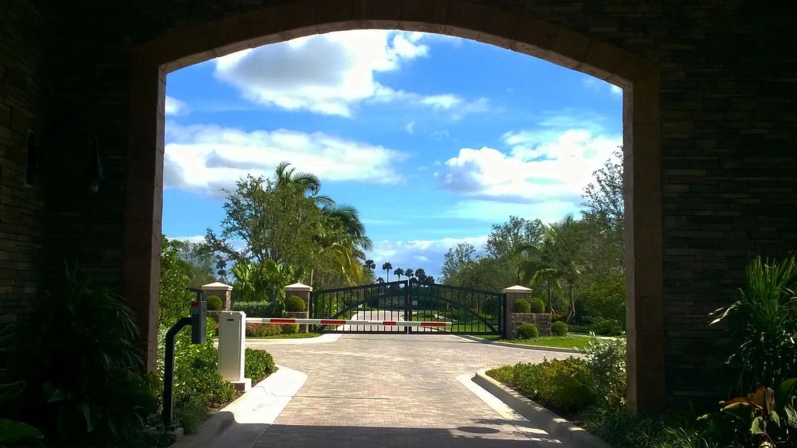 Sunlit stone driveway leading through an arched entrance to a gated garden under a blue sky with clouds