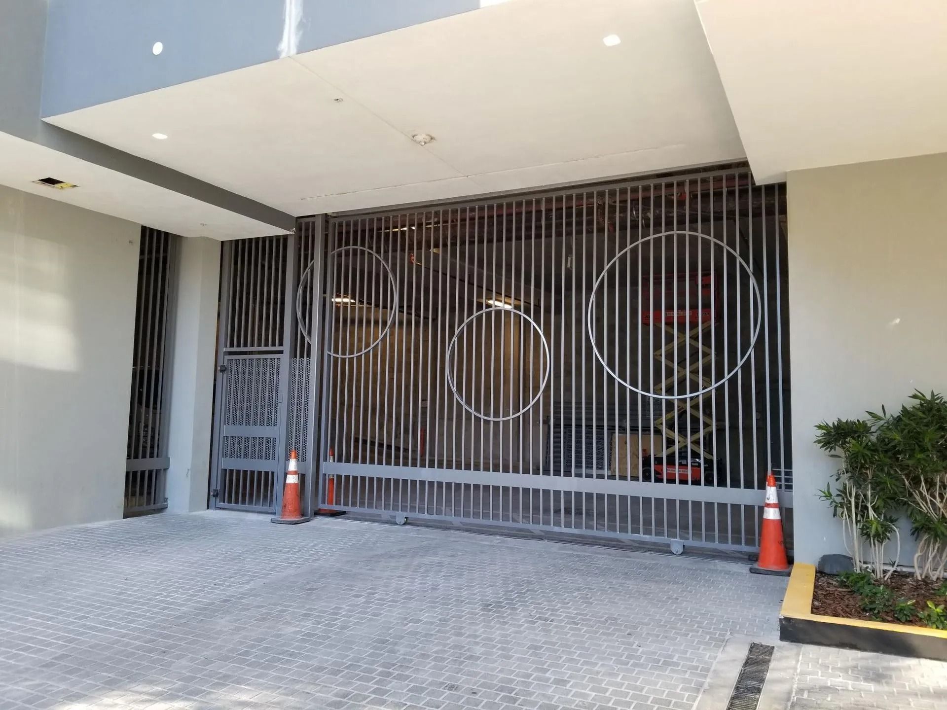 Modern house entrance with a tall metal gate, tiled driveway, and orange traffic cones at the sides
