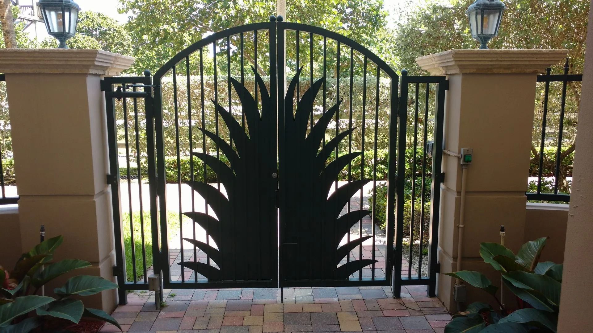 Black wrought-iron gate with a sunburst design between stone pillars and greenery beyond.