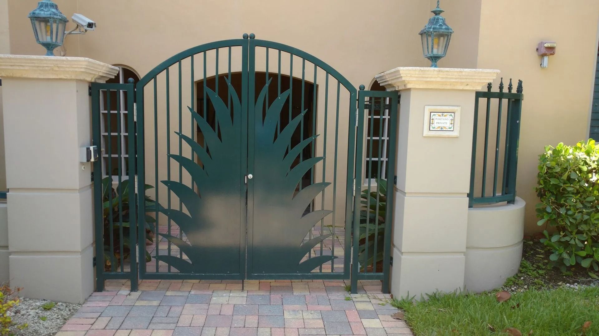 Dark green metal garden gate with decorative leaf design between stucco pillars and a paved walkway