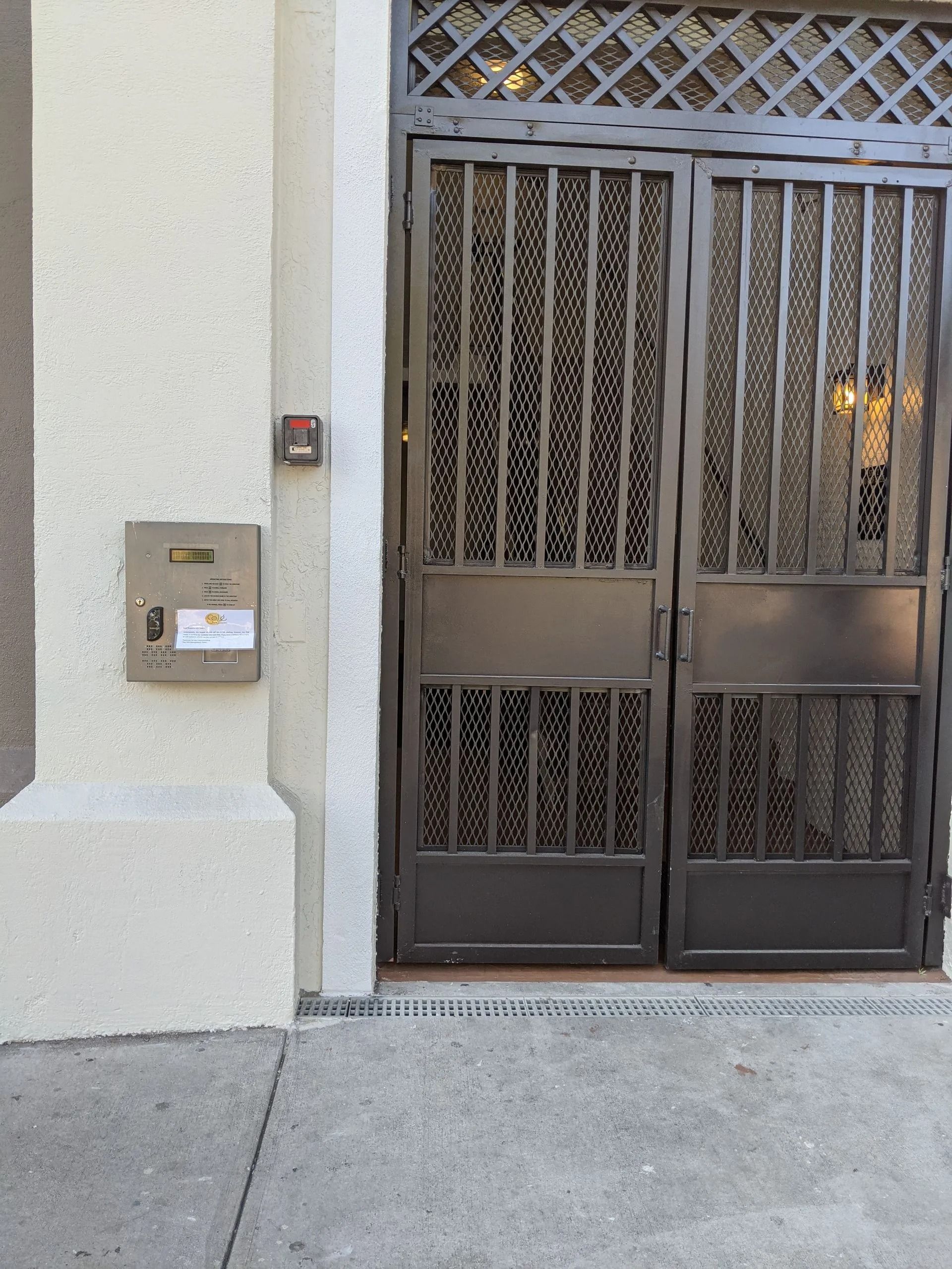 Metal gate entrance beside a beige wall with a wall-mounted mailbox and concrete sidewalk.