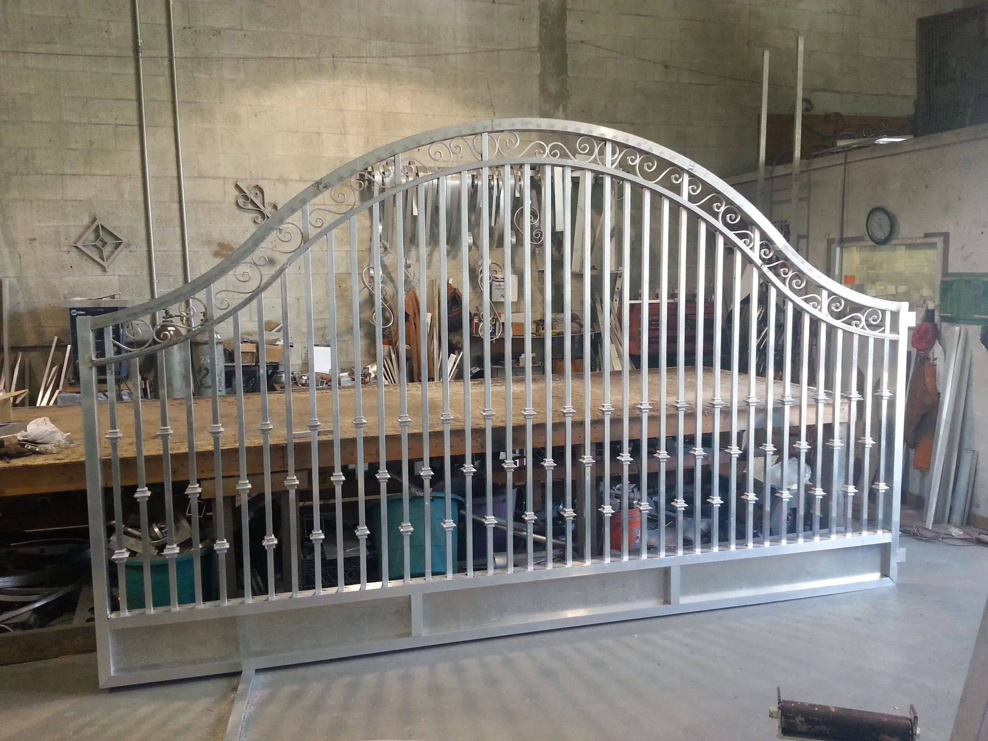 Silver metal gate with ornate wavy top, standing in a workshop against a concrete wall