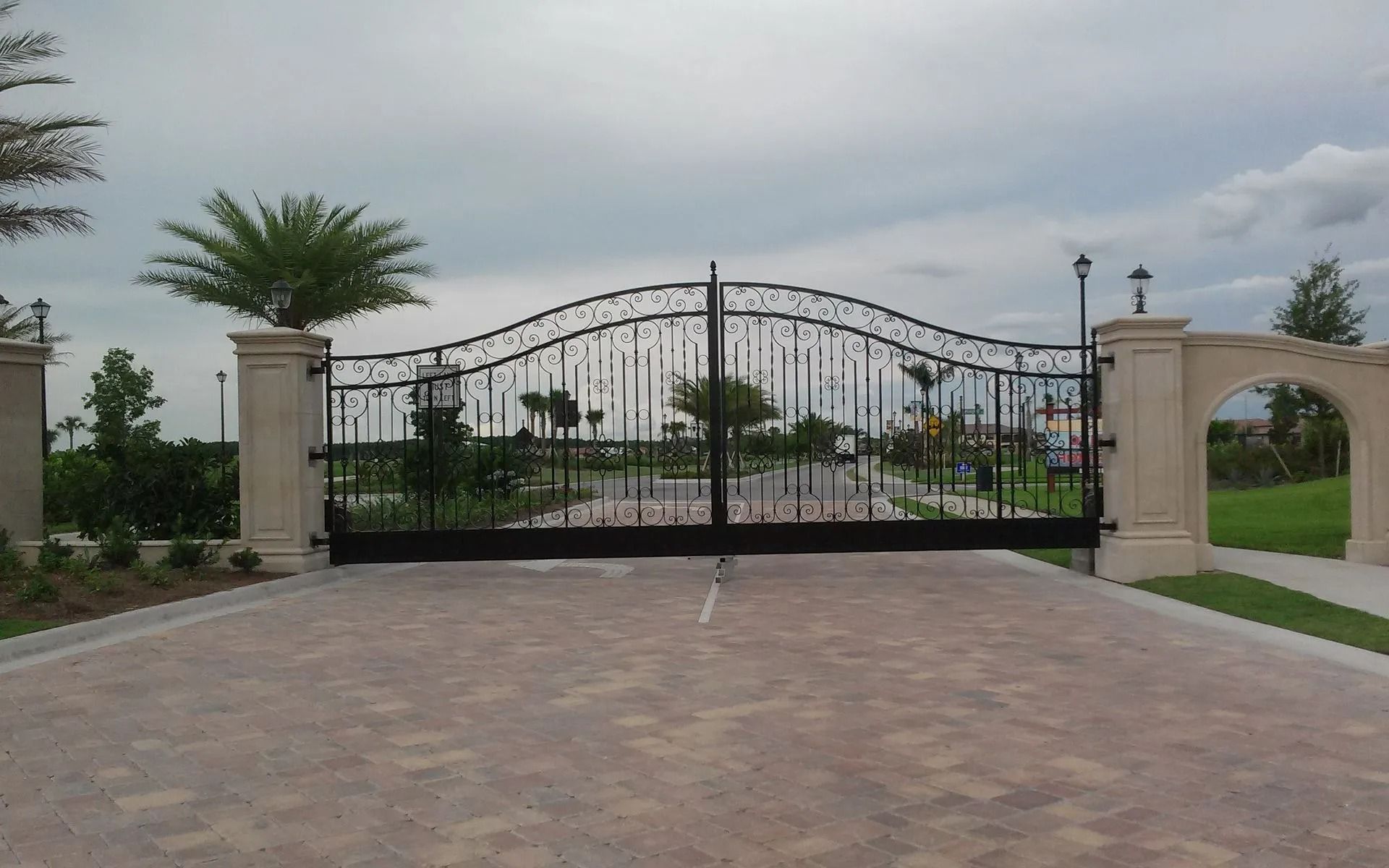 Wrought-iron driveway gate with stone pillars and palm trees under a cloudy sky