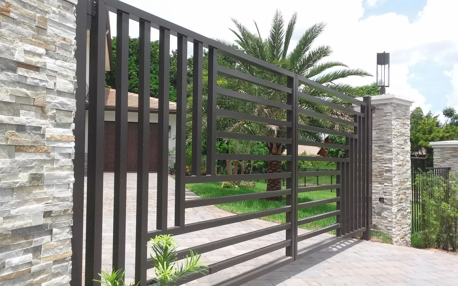 Modern metal driveway gate with stone pillars and tropical landscaping