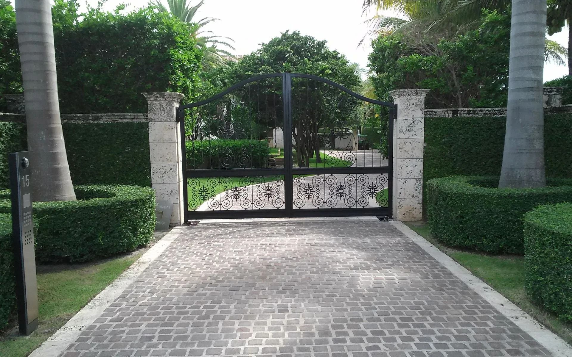 Paved driveway leading to a closed black gate between stone pillars, lined with palm trees and hedges.