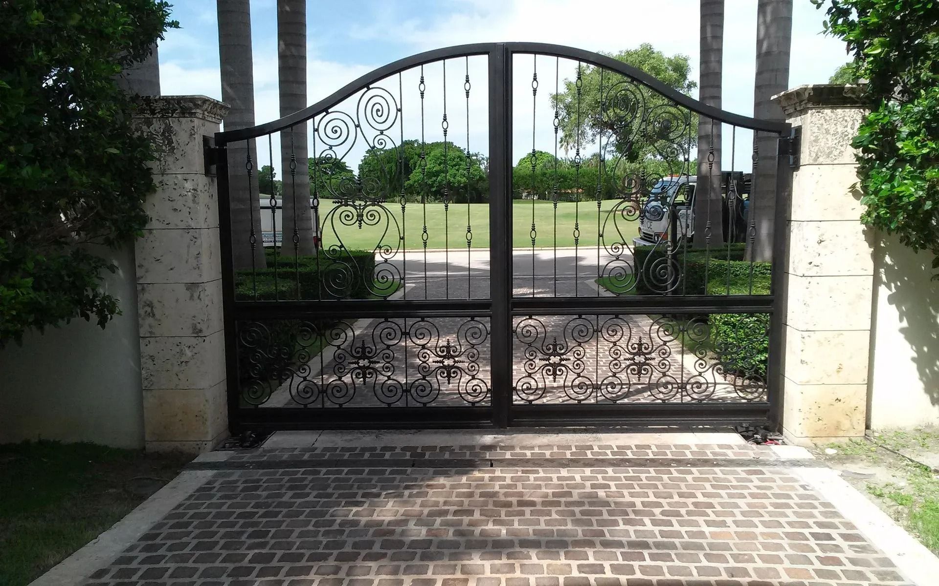 Black wrought-iron gate at the end of a brick driveway, opening to a sunny garden and street beyond.