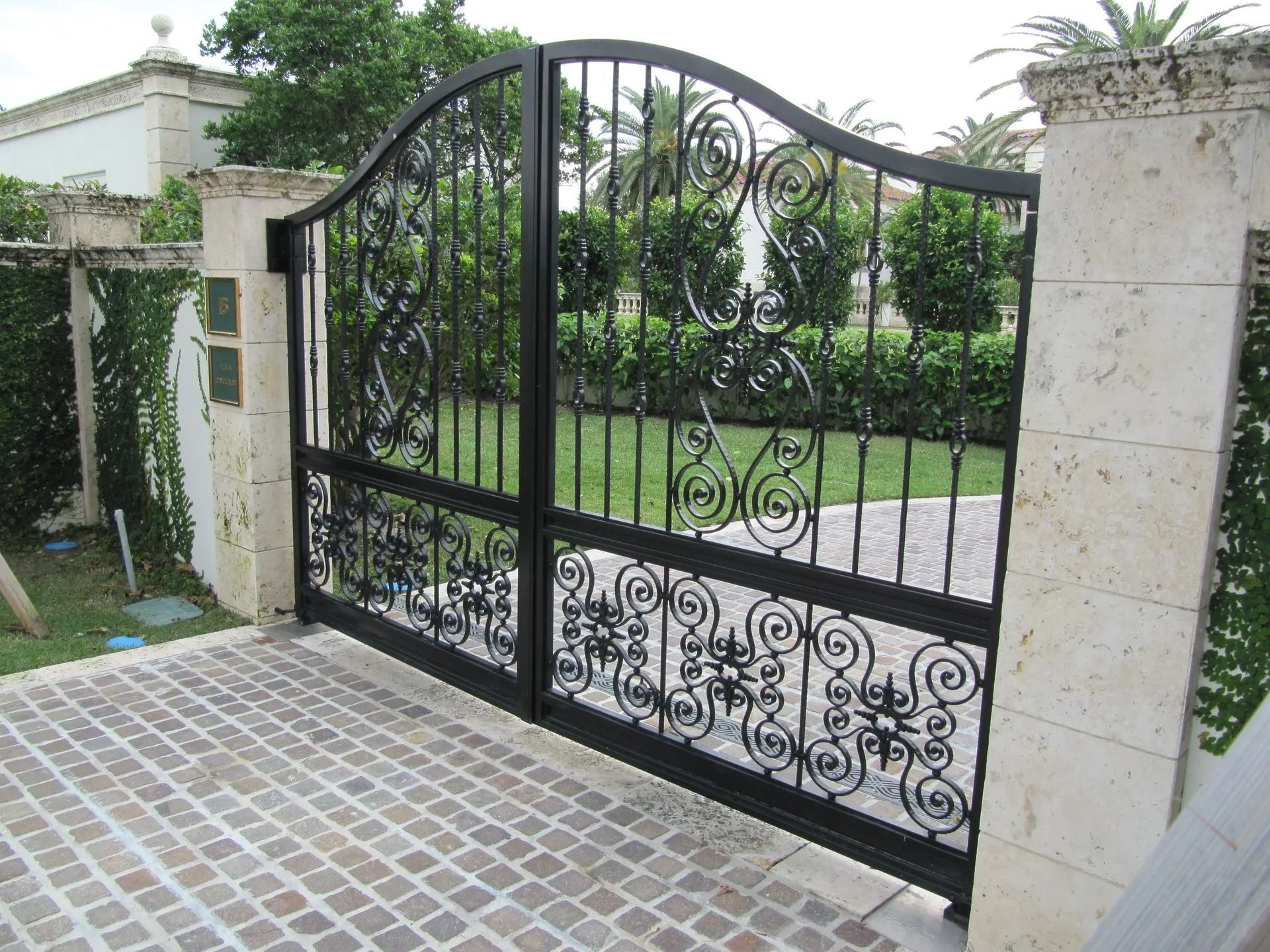 Black wrought-iron driveway gate between stone pillars at a landscaped entrance