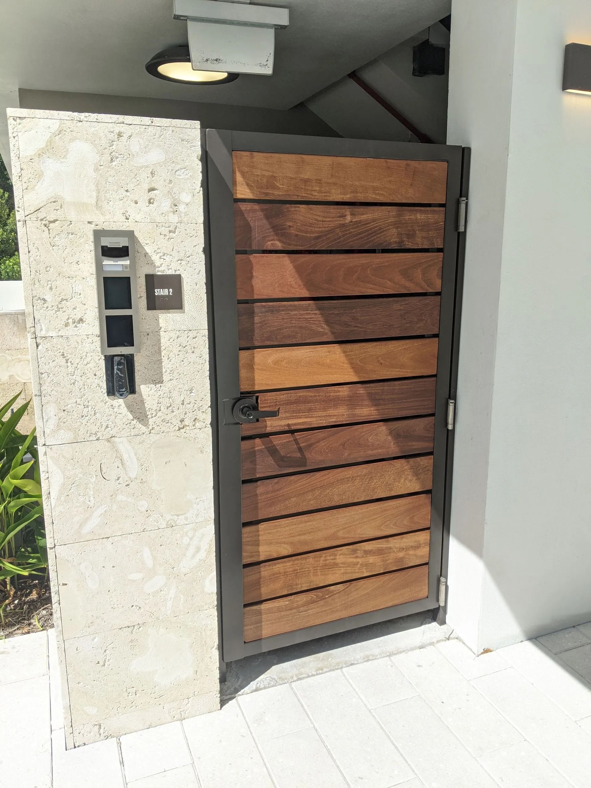 Front entry gate with wood slats, keypad intercom, and white stucco wall in bright sunlight