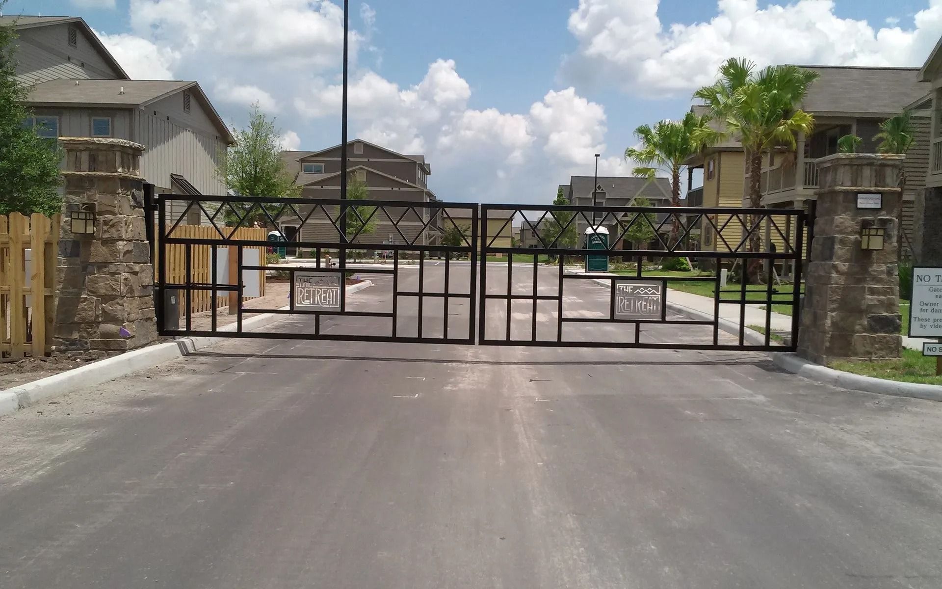 Gated entrance to a residential complex with stone pillars, metal gate, and road leading inside under a blue sky
