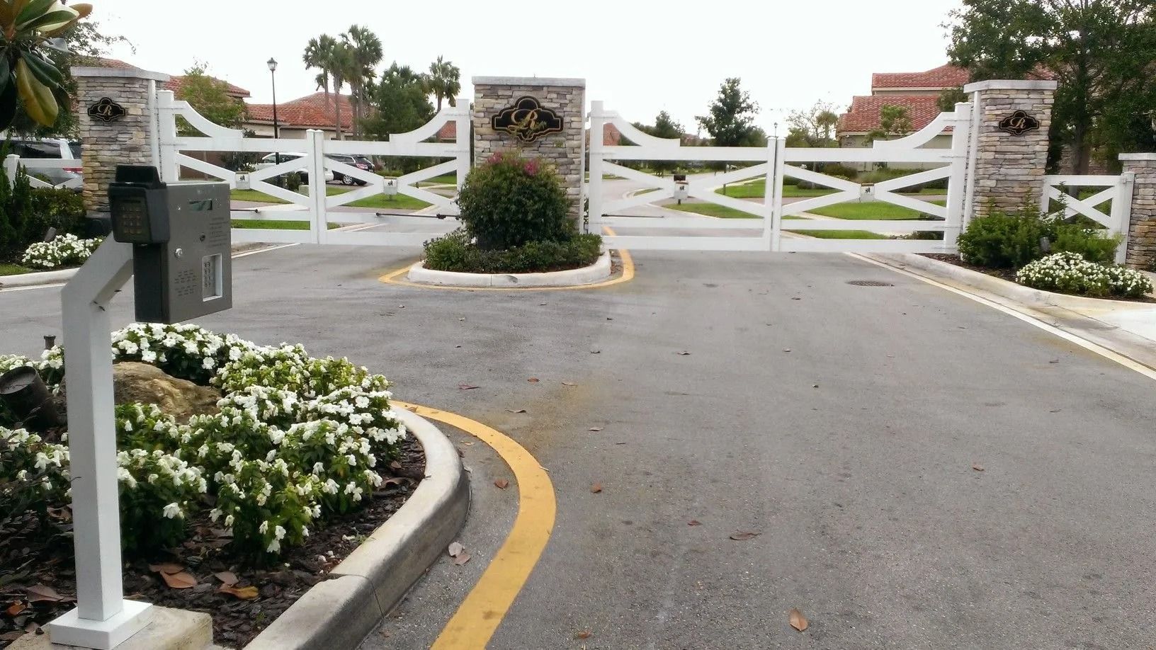 White gated driveway entrance with black mailbox, landscaped flower beds, and a curved asphalt road