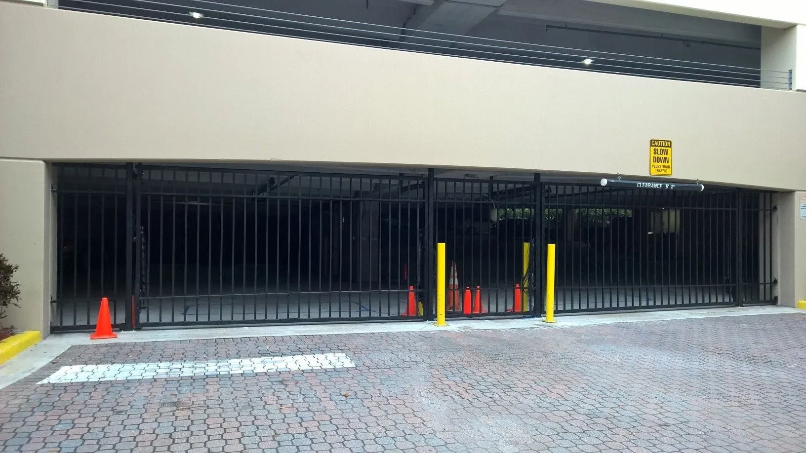 Gated parking garage entrance with black bars, yellow bollards, and orange traffic cones on a paved driveway