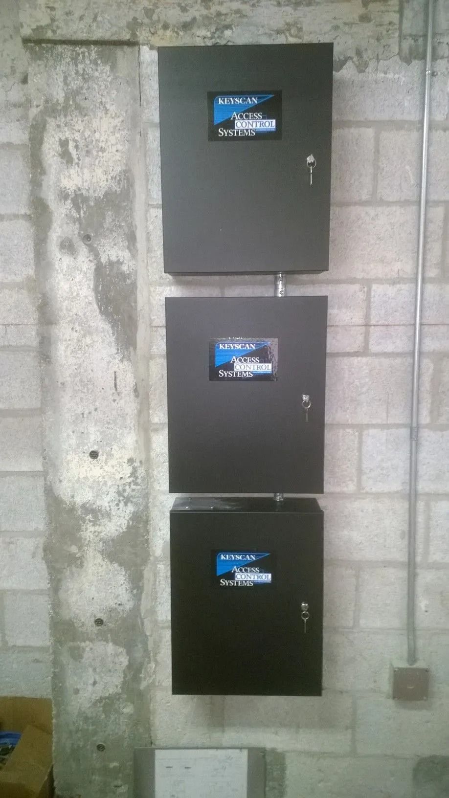 Three black electrical panels mounted vertically on a gray cinderblock wall.