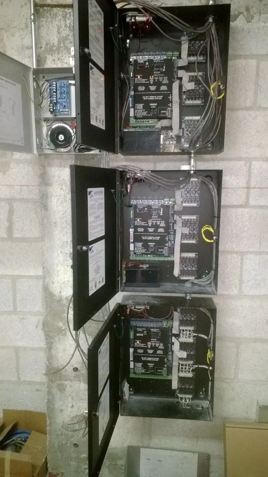 Three open black electrical control panels mounted on a cinderblock wall with wiring and labels visible