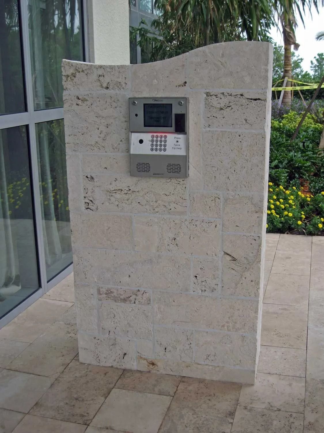 Stone outdoor intercom panel mounted on a beige wall beside glass doors and garden flowers