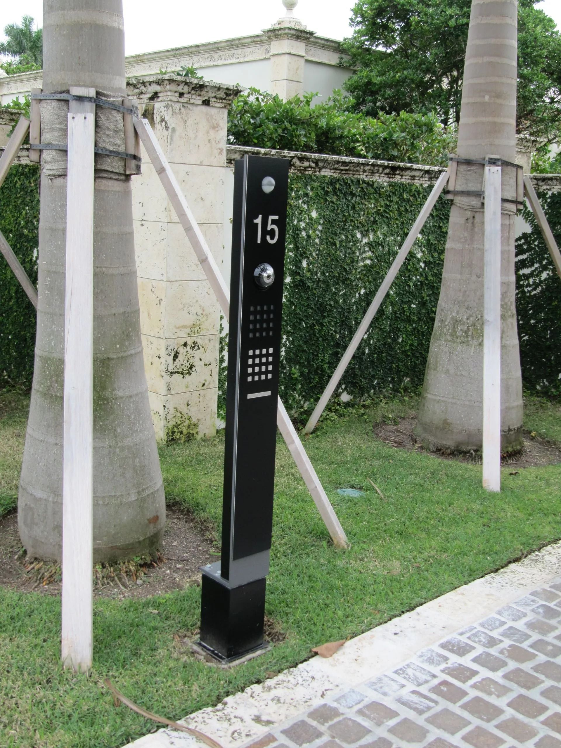 Black outdoor call button post labeled 15 beside palm trees and grass near a paved path