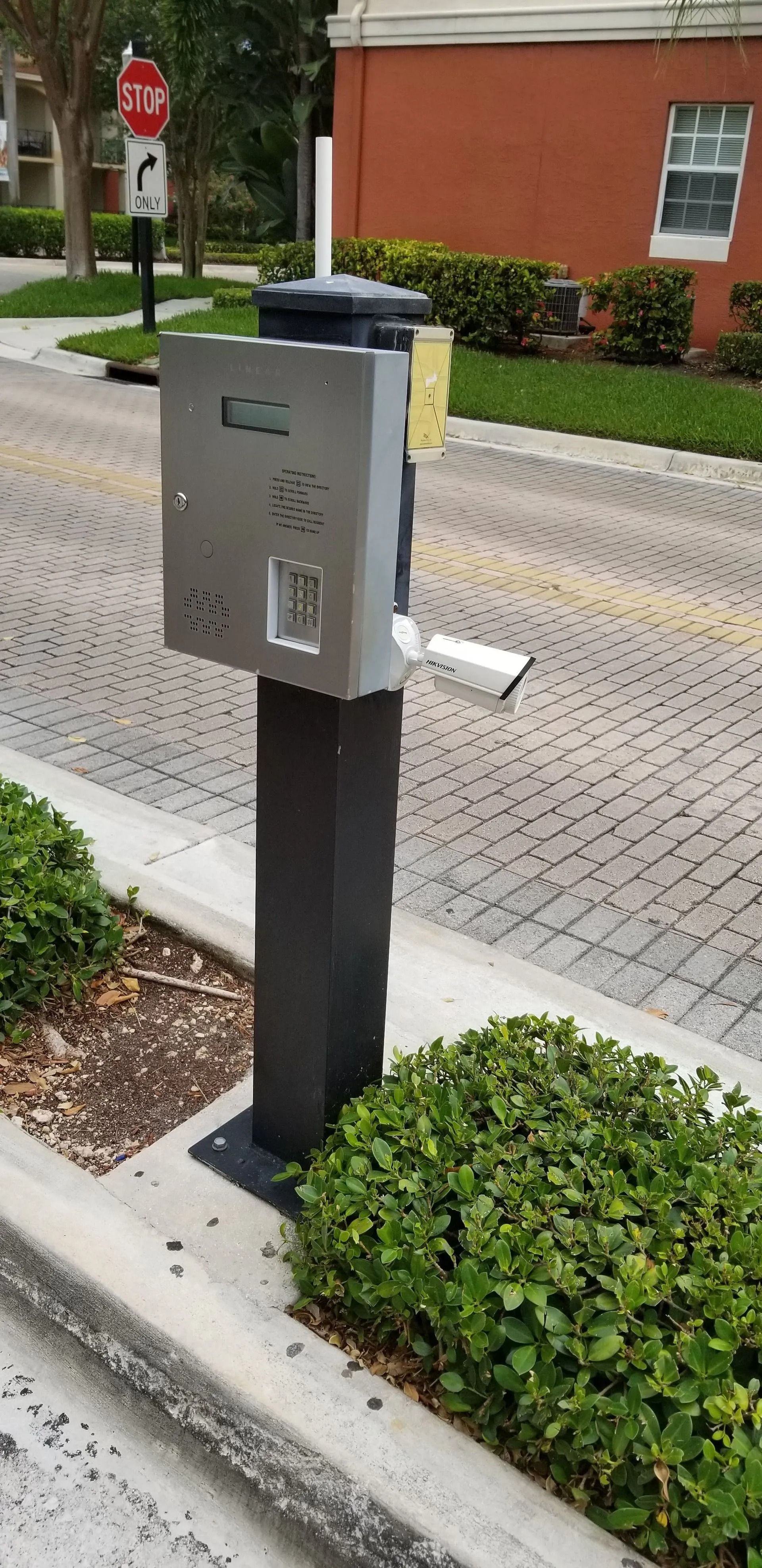 Gray roadside utility box on a post beside a brick building and sidewalk, with a stop sign in the background
