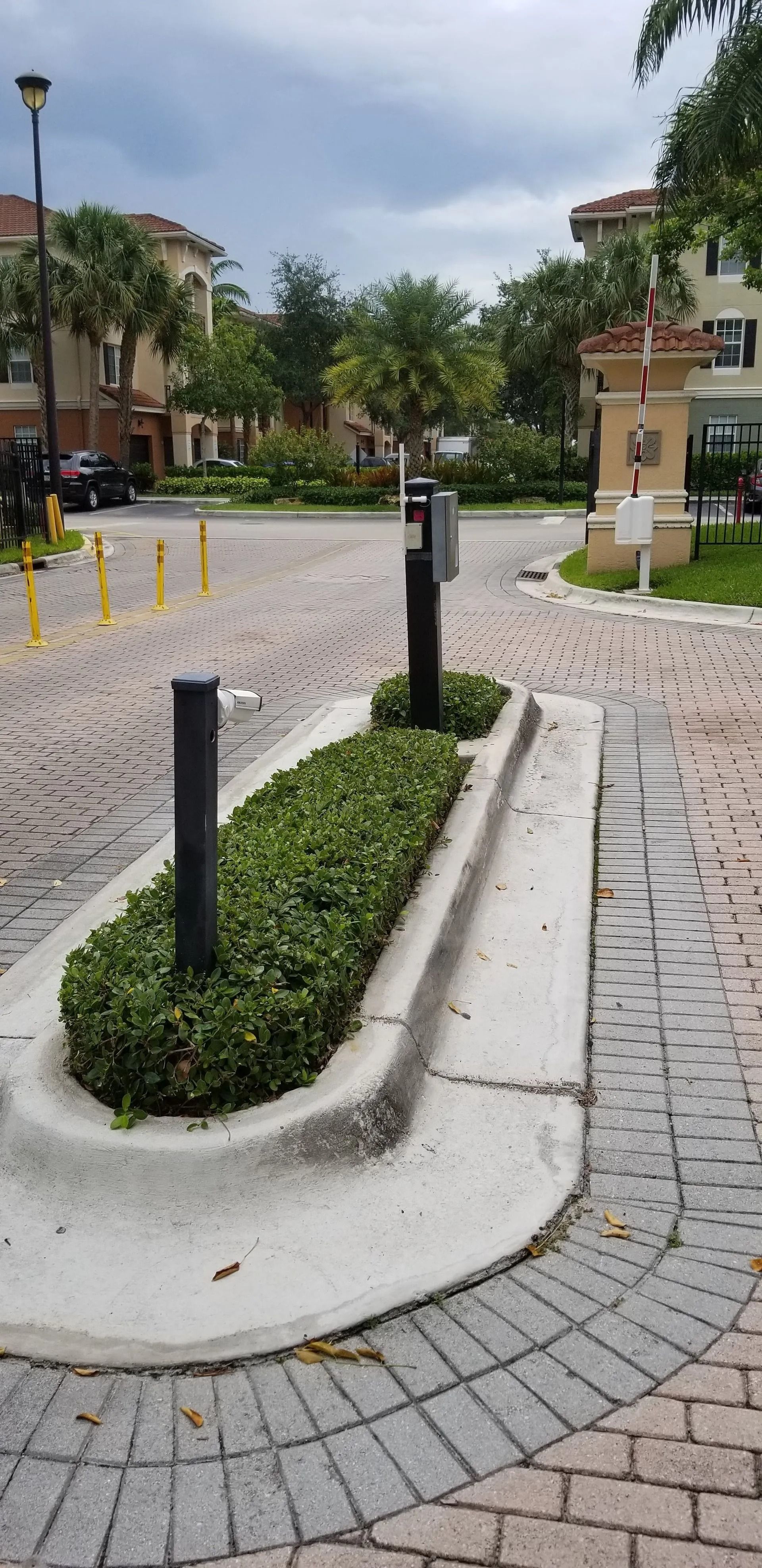 Curved landscaped pathway with black posts and yellow bollards in a cobblestone park, under cloudy skies