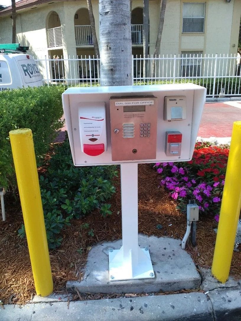 Outdoor emergency call box with red buttons and keypad on a white pedestal between yellow bollards.