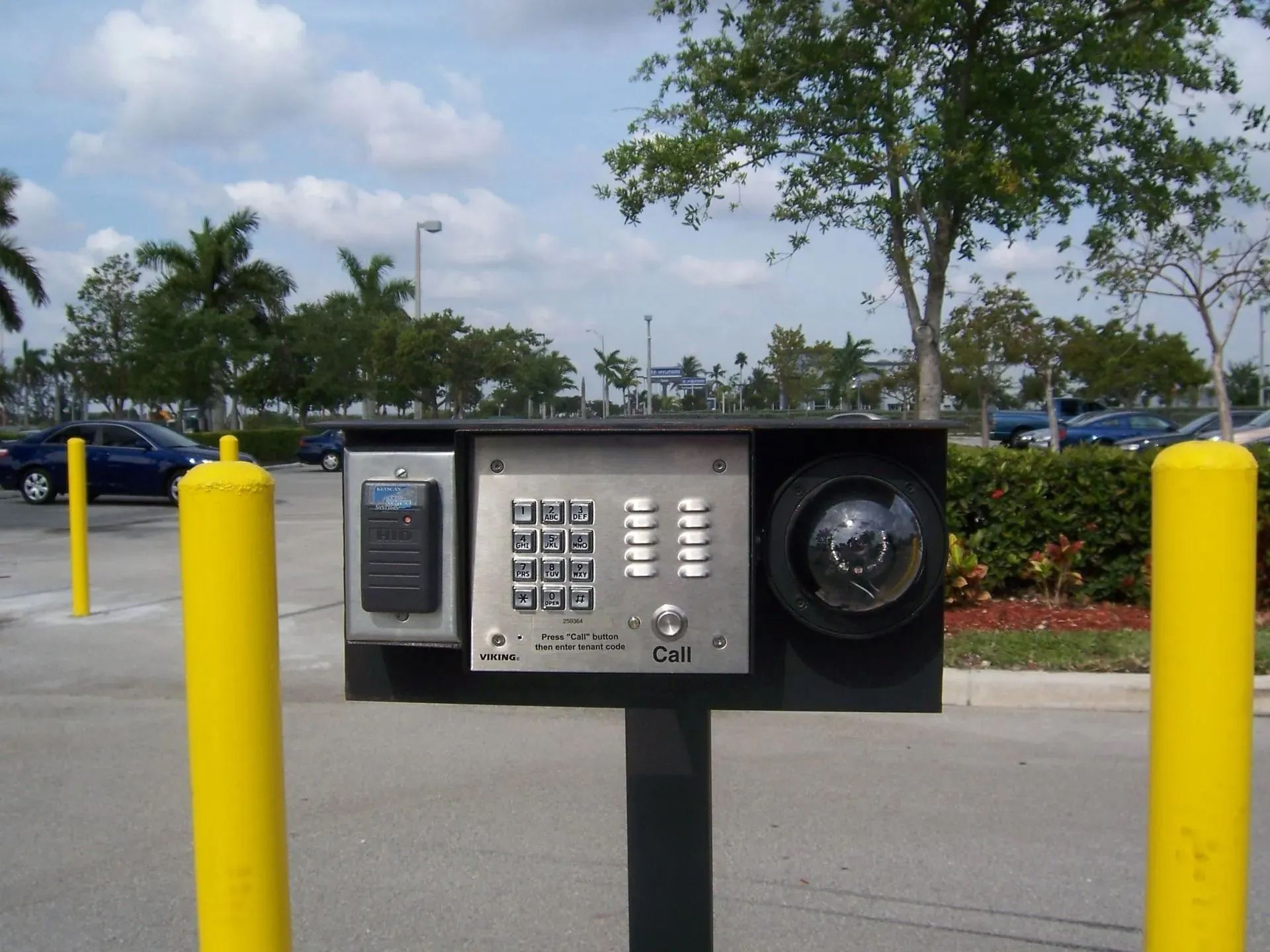 Outdoor parking lot payment kiosk with keypad and speaker, mounted on a post between yellow bollards.