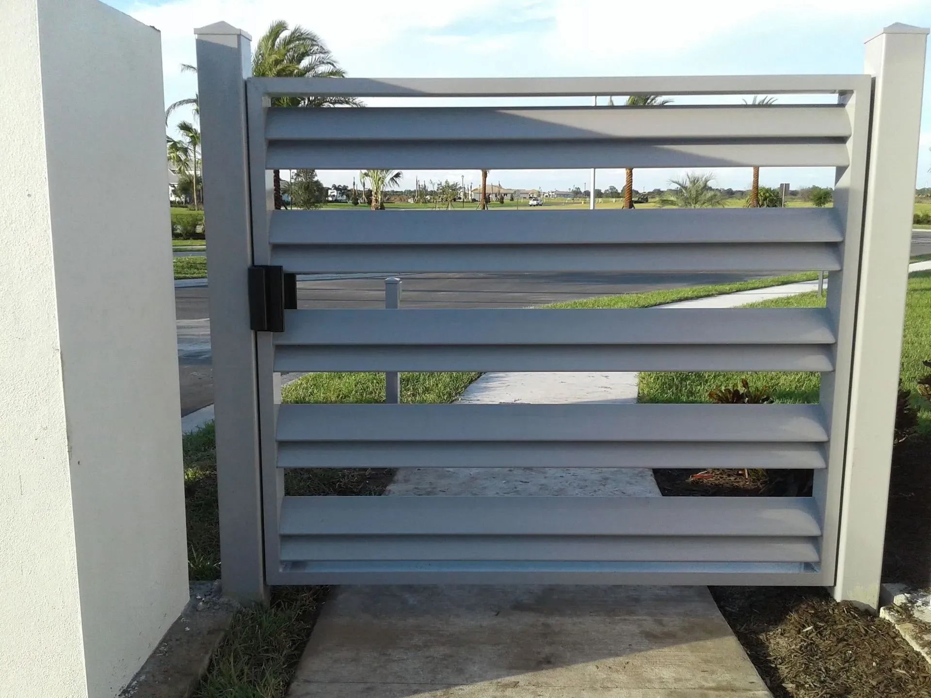 Gray slatted metal gate across a walkway beside a white wall, with greenery beyond.