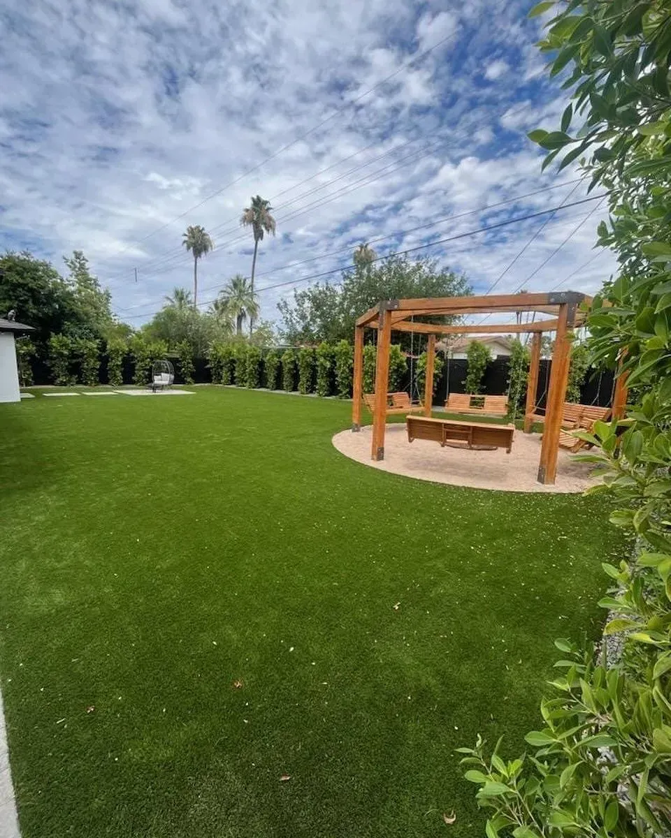 A grassy backyard with a wooden pergola, lush green lawn, trees, and a cloudy sky.