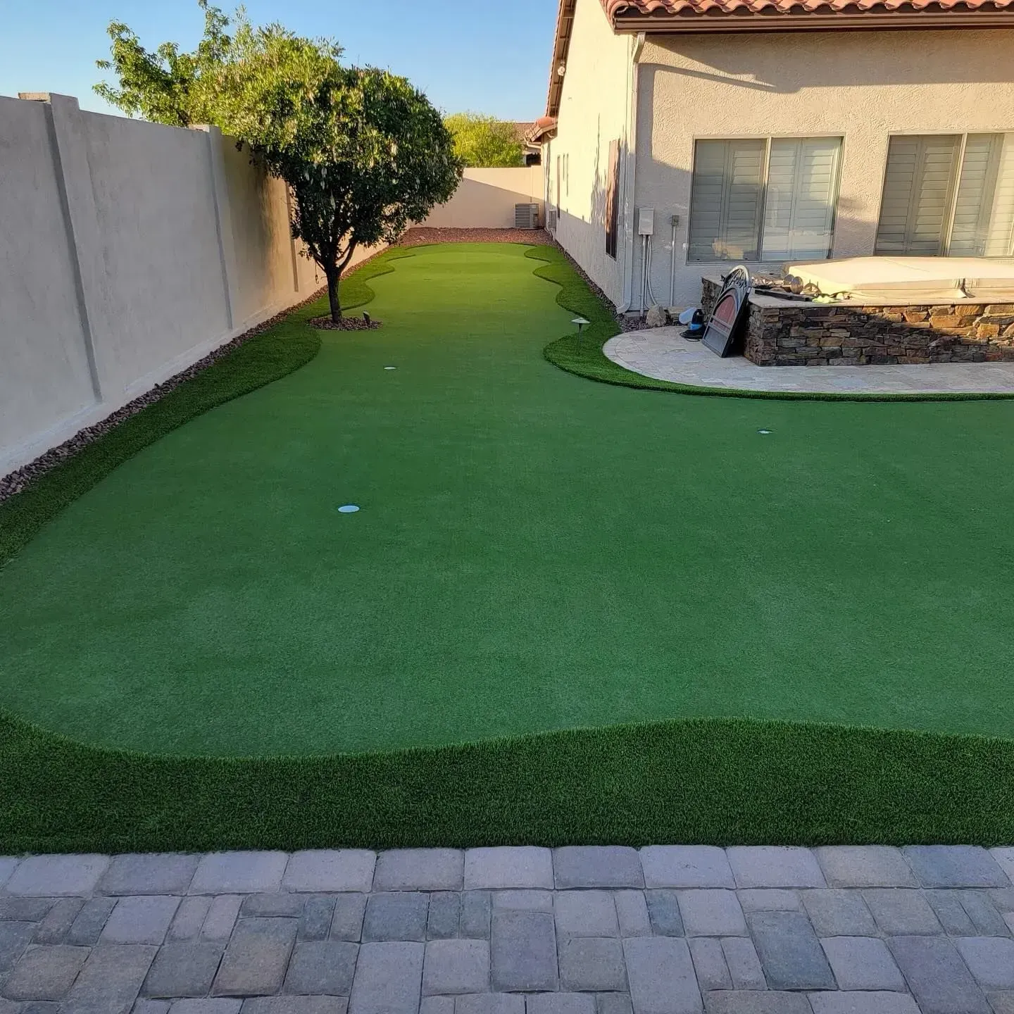 Backyard with putting green, artificial turf, and a small tree. Beige house and wall in background.