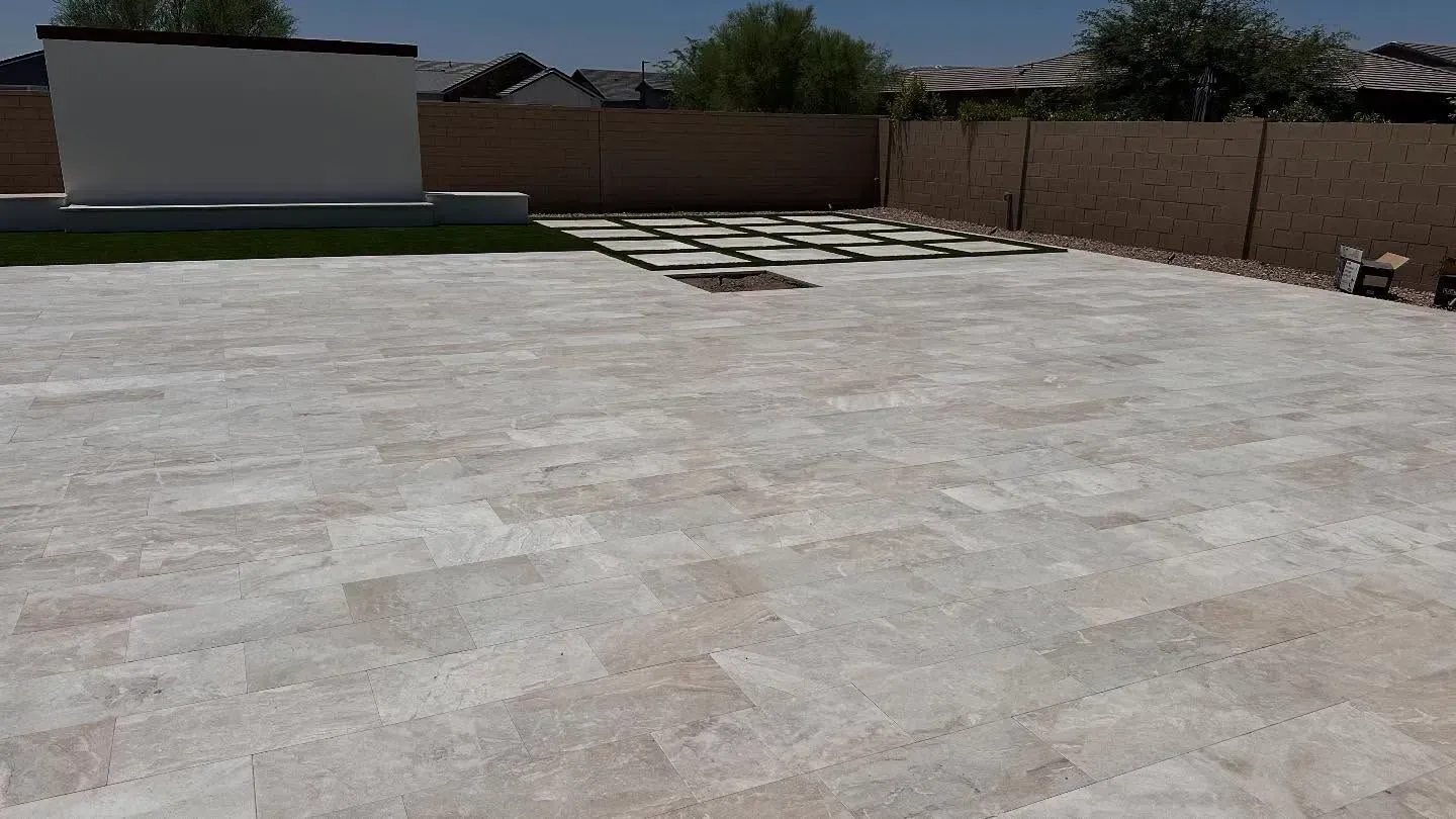 A backyard patio with tan paving stones and a raised rectangular area of green grass.